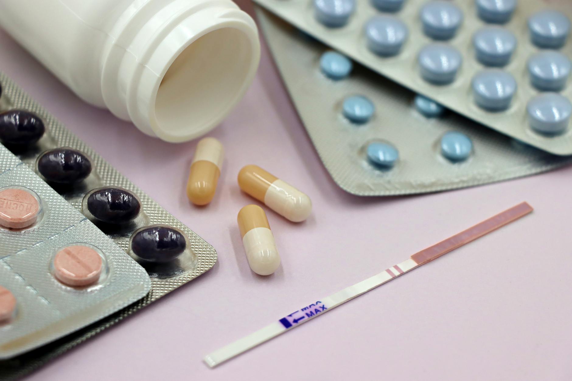 Close-up of various medications including capsules, pills, and an ovulation test strip on a pink background.
