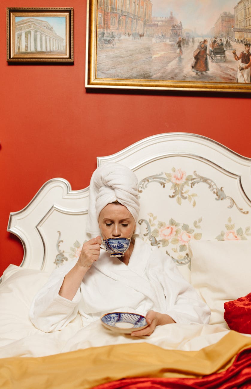A woman in a white bathrobe and a towel on her head sits in bed, sipping tea from a decorative cup, with a bowl in her hand and vintage artwork on the wall behind her.